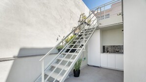 Un escalier blanc avec des rampes en métal menant à un balcon, le tout entouré d’un mur blanc et agrémenté d’une plante en pot.