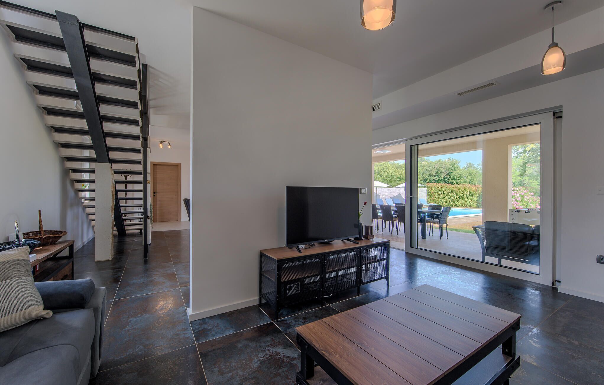 A modern living room with a TV, a wooden coffee table, and a sofa. There's a staircase on the left and a sliding glass door leading to an outdoor patio.