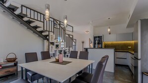 A modern kitchen with a central island, a dining area with a white table and chairs, and a staircase with a railing.