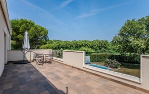 Ein Balkon mit Fliesenboden, einem Tisch mit Stühlen, einem Sonnenschirm und Blick auf einen Swimmingpool und Grünflächen.