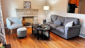 A living room with a stone fireplace, grey sofa, armchair, and coffee table. - The Shaded Retreat (Louisville)