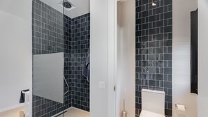 Ein modernes Badezimmer mit dunkel gefliesten Wänden, einer Duschkabine aus Glas, einer weißen Toilette und einem kleinen Mülleimer.