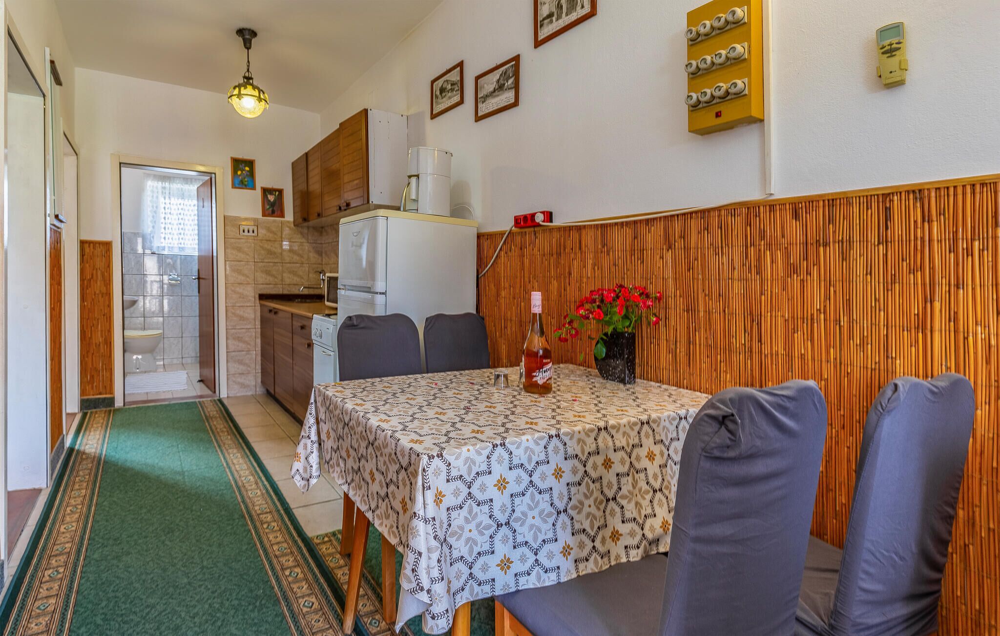A compact kitchen with a refrigerator, microwave, and wooden cabinets. A dining area with a table and chairs is visible, along with a bamboo wall panel and a small potted plant.