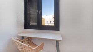 A room with a white table, a wicker chair, a dark-framed window, and a view of buildings outside.