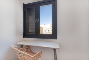Habitació amb taula blanca, cadira de vimet, finestra amb marc fosc i vista a edificis.
