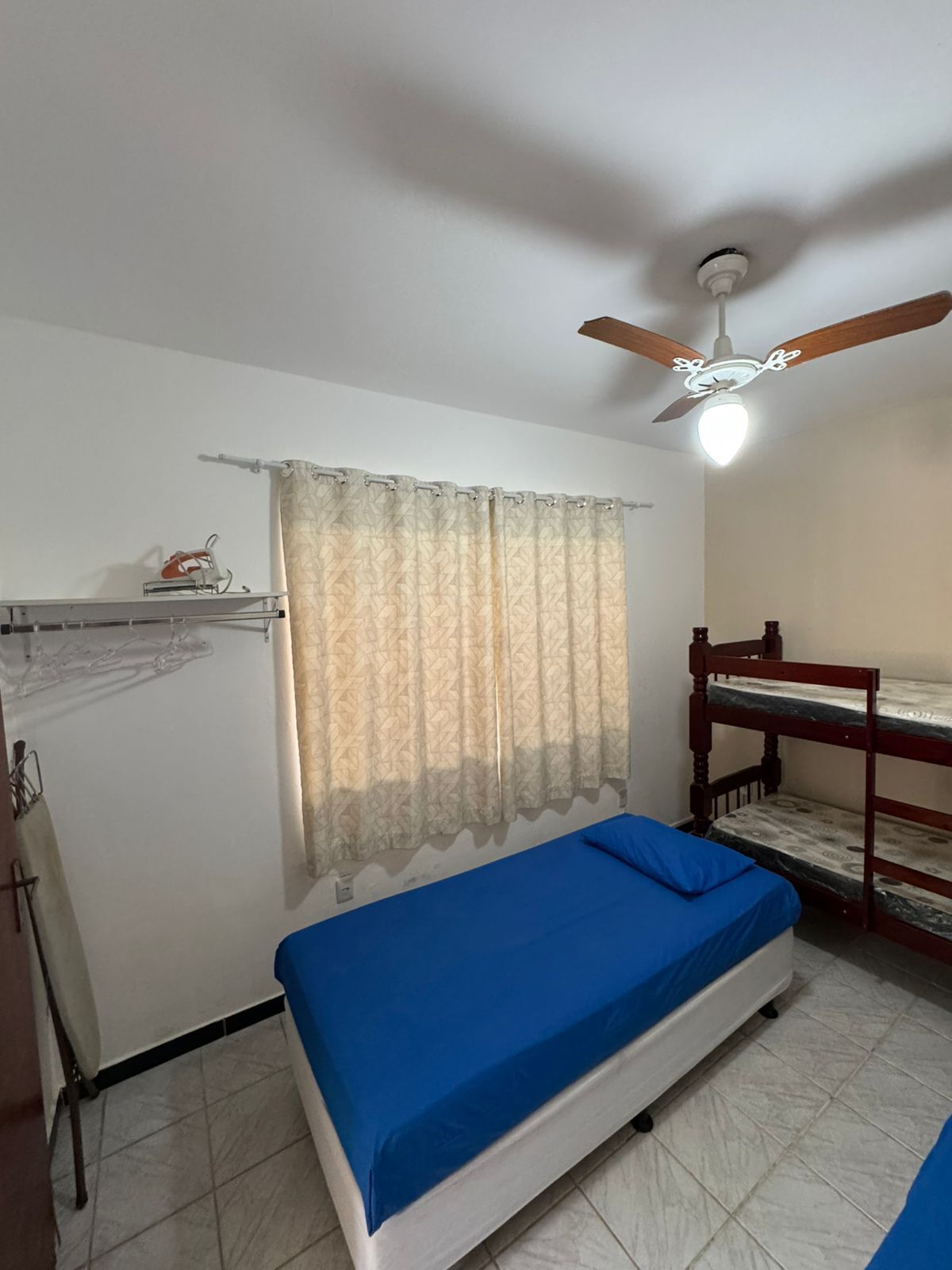 Quarto com uma cama beliche, ventoinha no teto, janela com cortinas estampadas e cama com colchão azul.