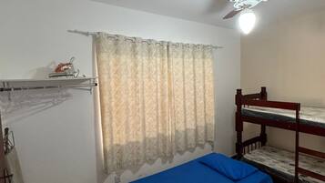 Quarto com uma cama beliche, ventoinha no teto, janela com cortinas estampadas e cama com colchão azul.