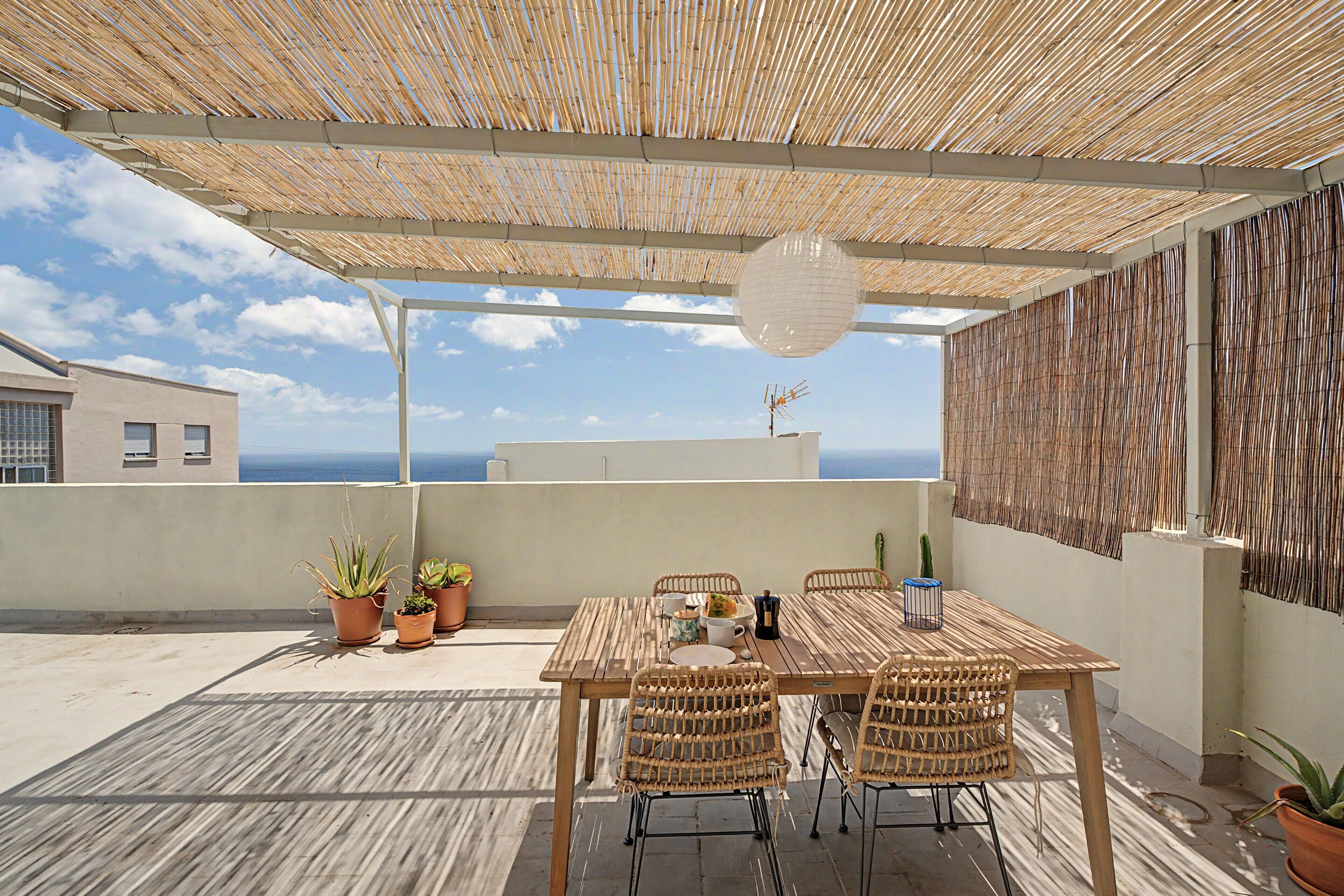 Eine Terrassengruppe im Freien mit Holztisch, Korbstühlen, Topfpflanzen und Blick aufs Meer.