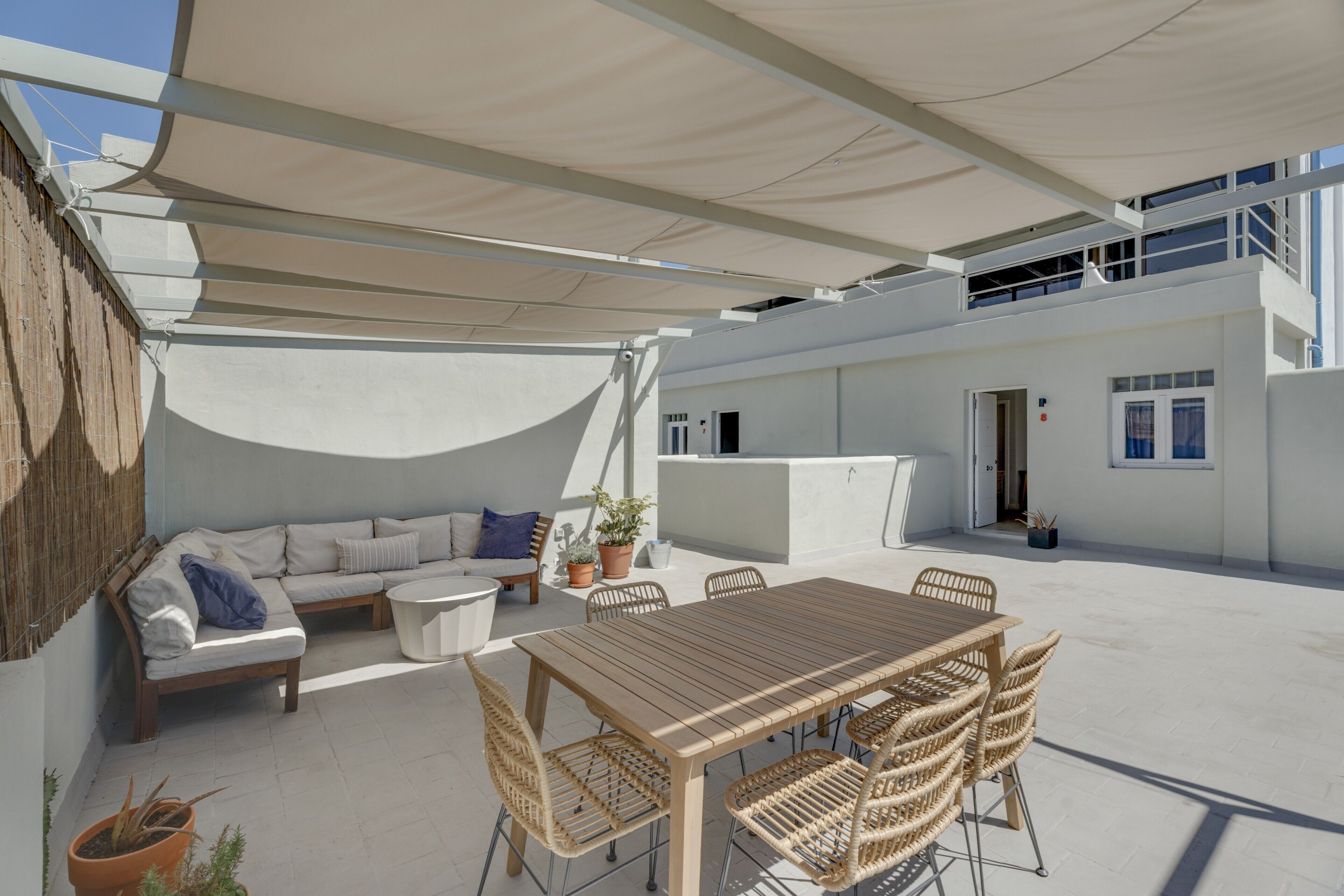 Una terraza en la azotea con una mesa y sillas de madera, un sofá, plantas en macetas y un edificio blanco al fondo.