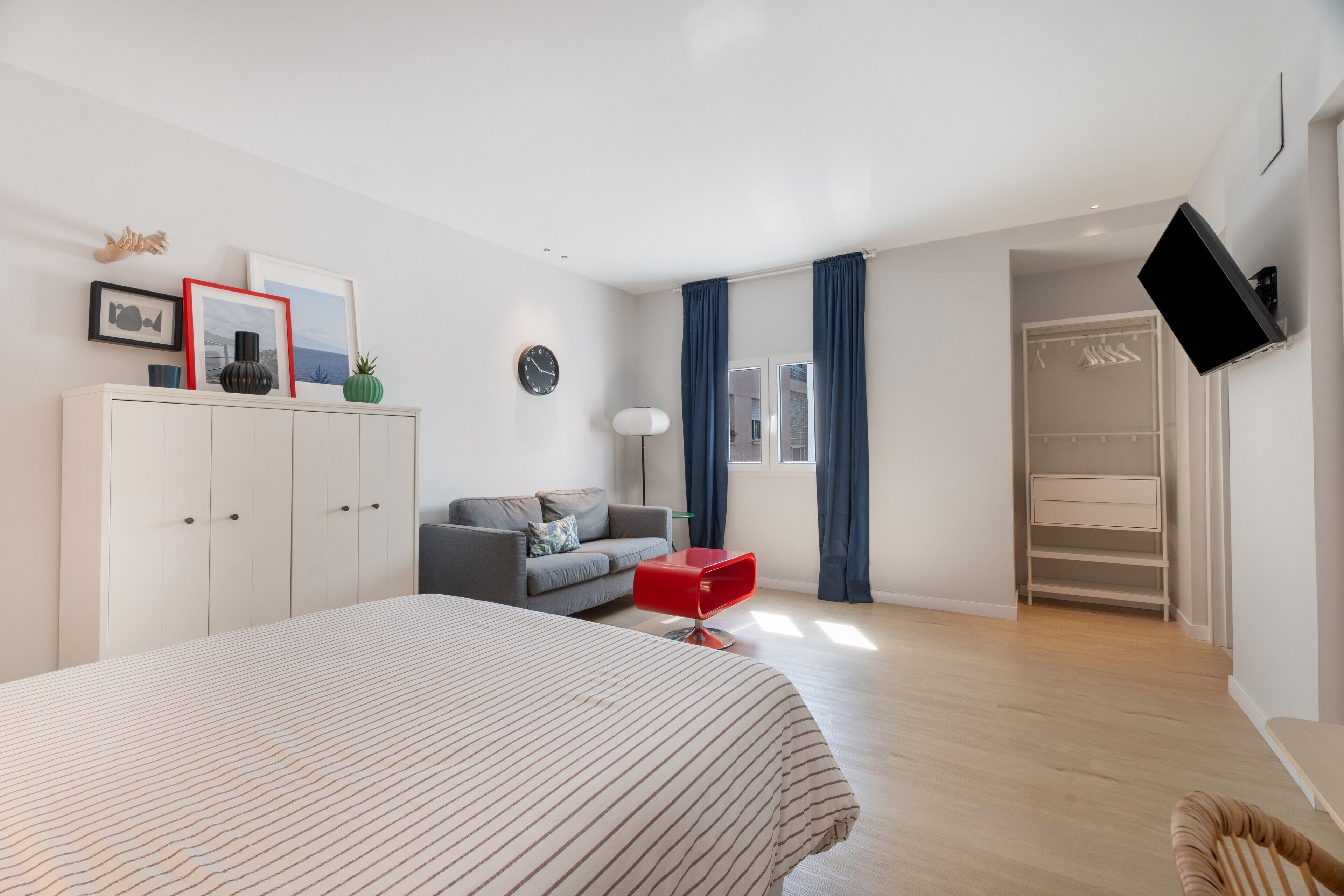 Un dormitorio moderno con una cama grande, un sofá gris, una silla roja, un armario, un televisor y un ropero.