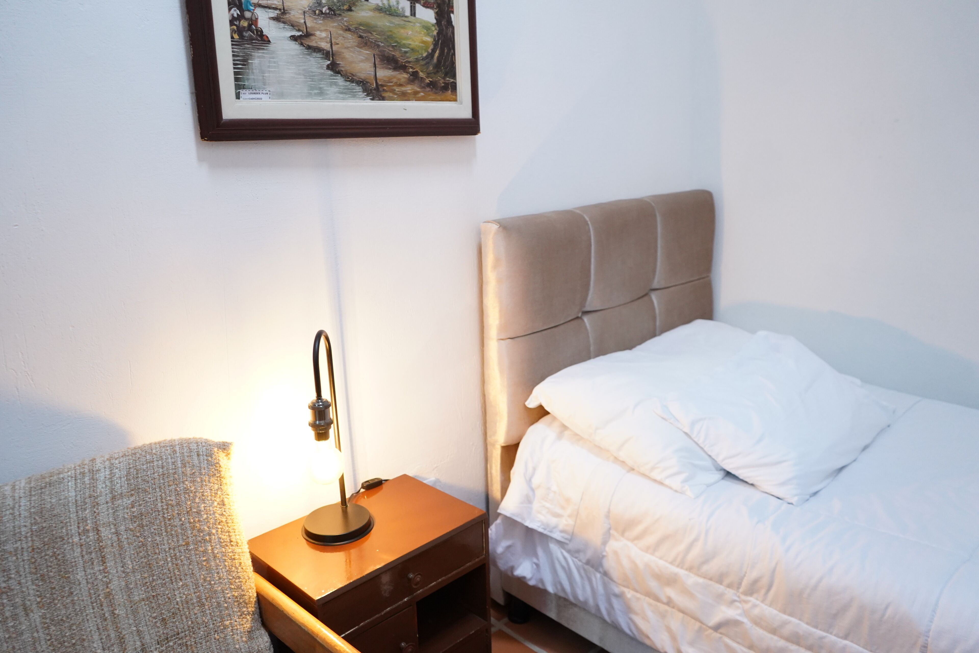 A neatly made bed with white bedding, a bedside table with a lamp, and a framed picture on the wall.