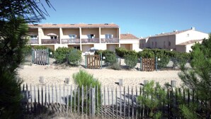 A multi-story building with balconies, surrounded by a sandy area with sparse vegetation.
