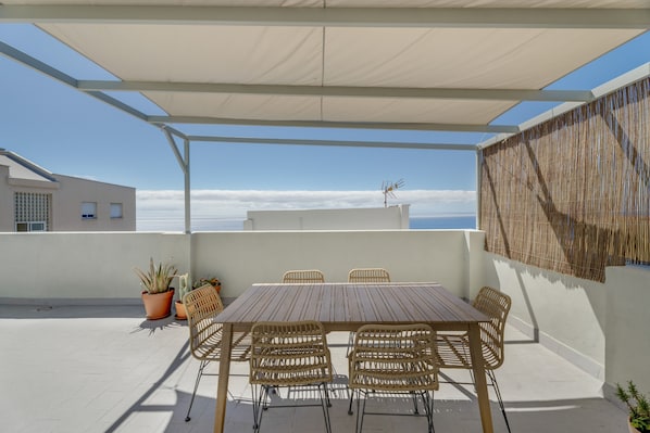 A covered outdoor patio with a wooden table and wicker chairs, potted plants, and a view of the sky.