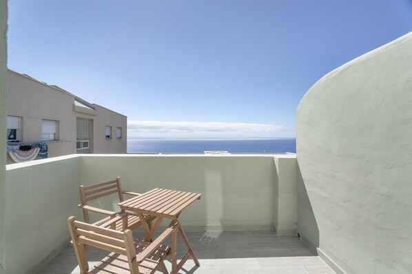 A balcony with a wooden table and chairs, a view of the sea, and a building with a light-colored exterior.