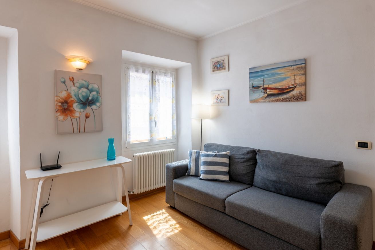 Ein Wohnzimmer mit grauem Sofa, einem weißen Beistelltisch, einem Blumenbild, einer Lampe und einem Fenster mit Vorhängen.