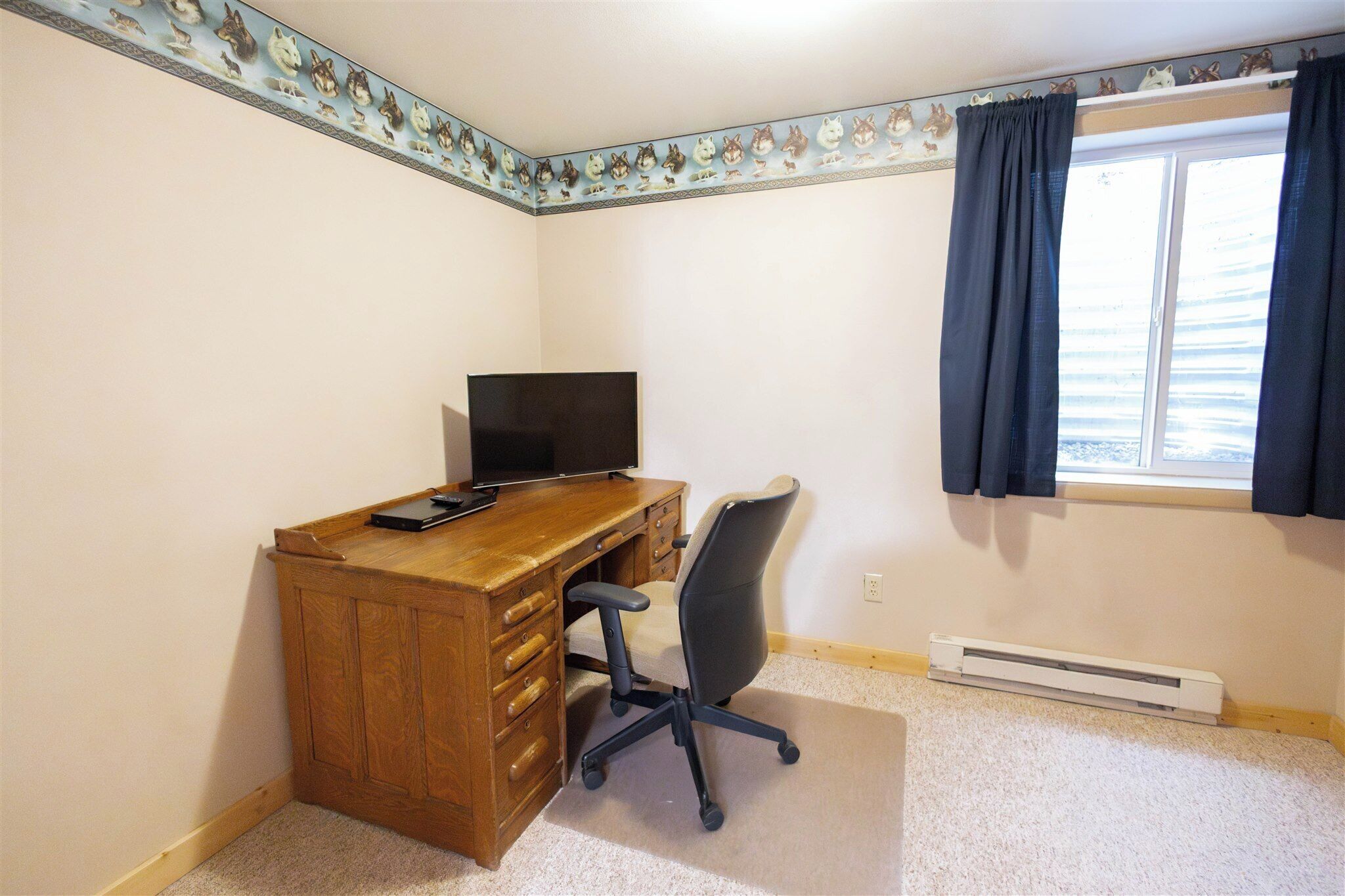 Une pièce avec un bureau en bois, un fauteuil de bureau noir, un téléviseur et une fenêtre avec des rideaux foncés.