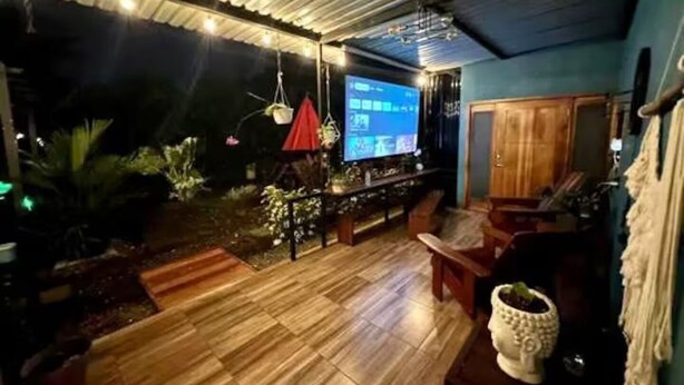An outdoor patio area with a wooden floor, a seating arrangement, hanging plants, and a television screen.