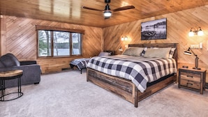 A cozy bedroom with a large bed, a desk, a chair, and a window overlooking a lake.