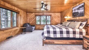 A cozy cabin bedroom with a bed, a chair, and a window overlooking a forest.