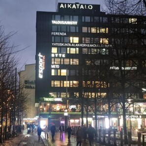 A building with multiple signs including 'AIKATALO', 'TALOUSJURISTIT', 'FERENDA', 'YHTEISKUNTA-ALAN KORKEAKOULU', 'STTK', 'METO', and 'FRIENDS & DRUGS'. - AikaTalo Hostel (Helsinki)