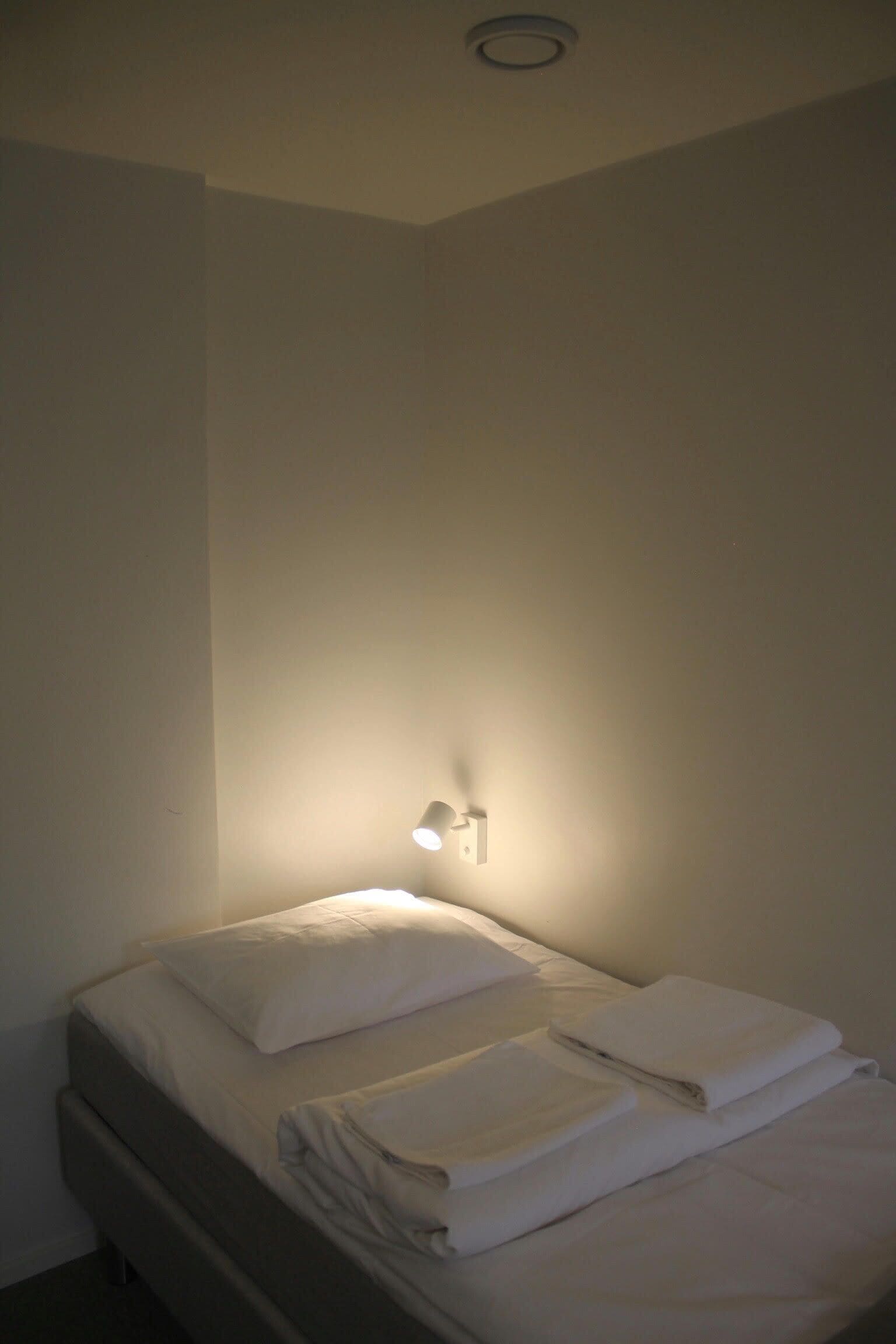 Una cama individual con sábanas y almohadas blancas, una lámpara de pared y una rejilla de ventilación en el techo.