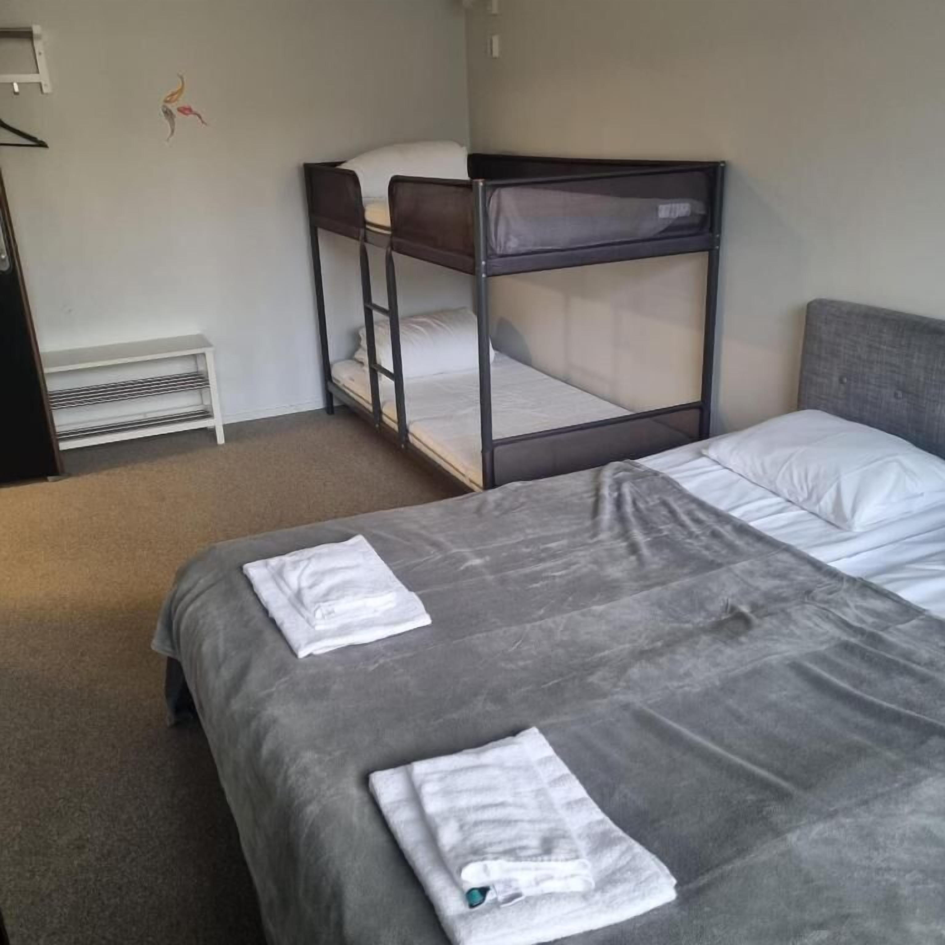 A room with a bunk bed and a single bed, both with white linens. There is a small white shelf unit against the wall and a radiator below it.