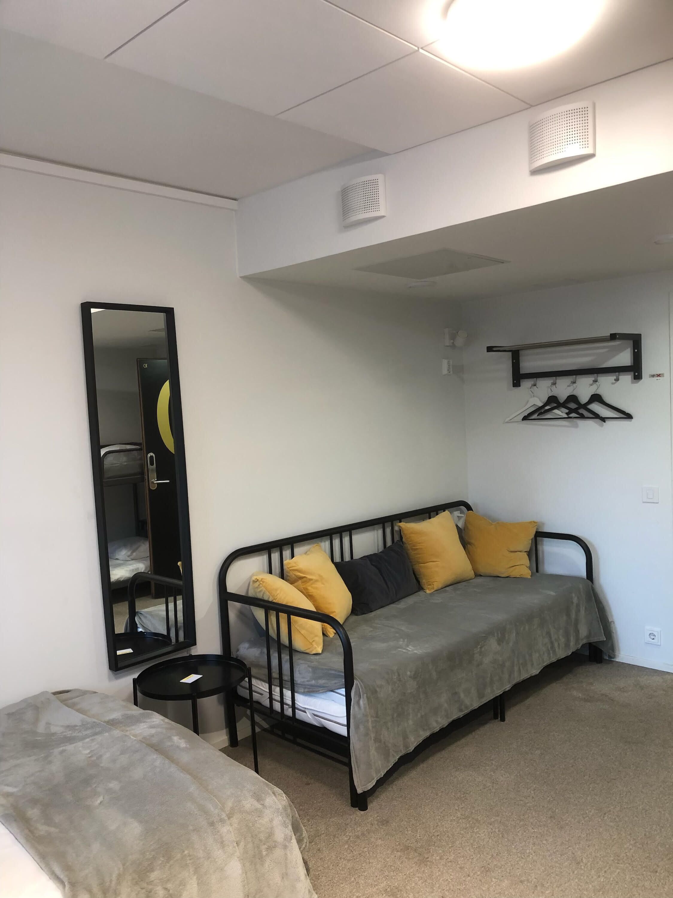 Um quarto pequeno e bem arrumado com uma cama, um sofá, um espelho e um cabideiro.