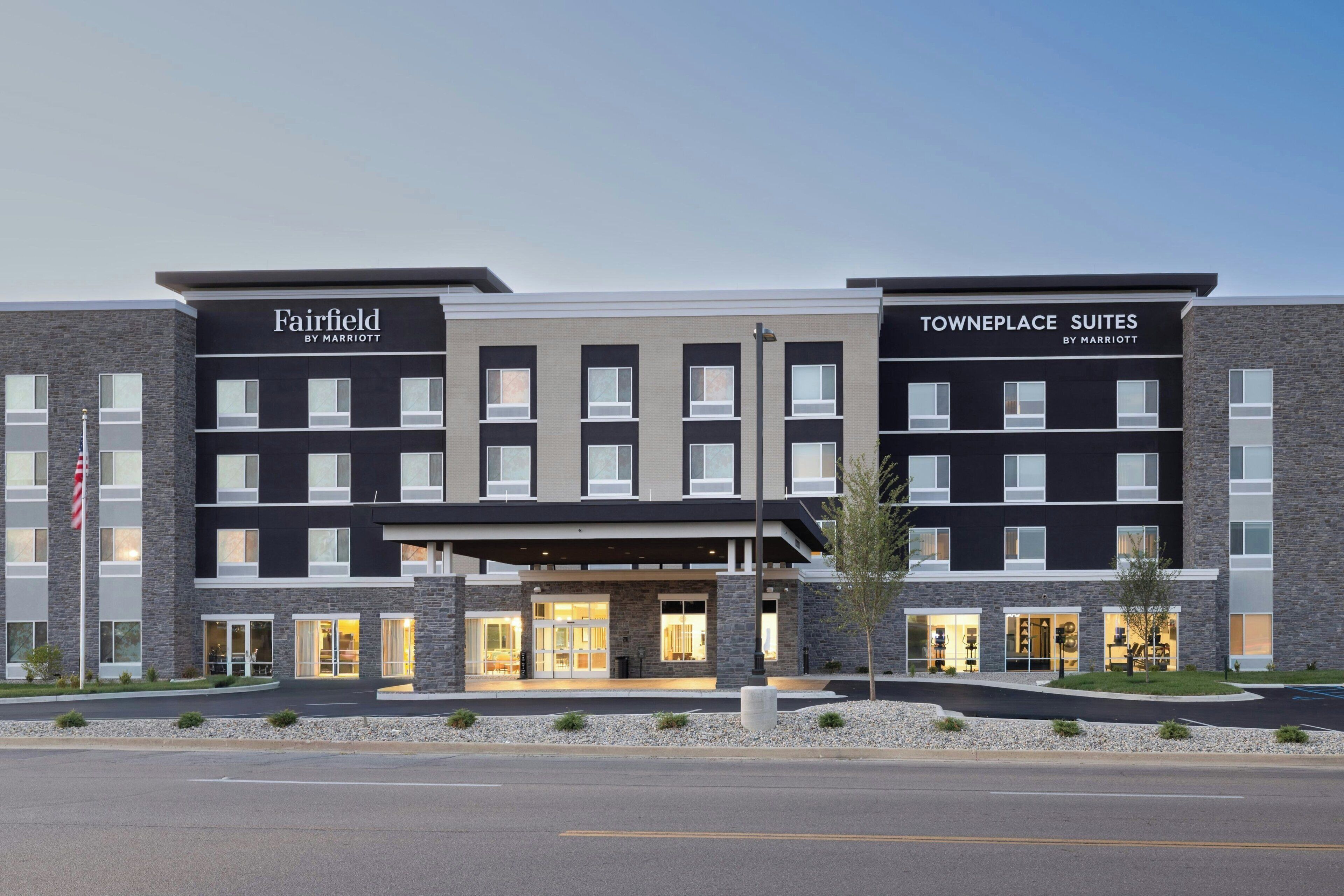 Un hotel moderno con señalización de Fairfield y Towneplace Suites by Marriott, que presenta una fachada simétrica con elementos de piedra y vidrio, y una entrada central con puertas de cristal.