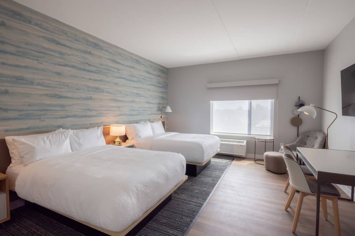 A modern hotel room with two beds, a desk, and a large window.