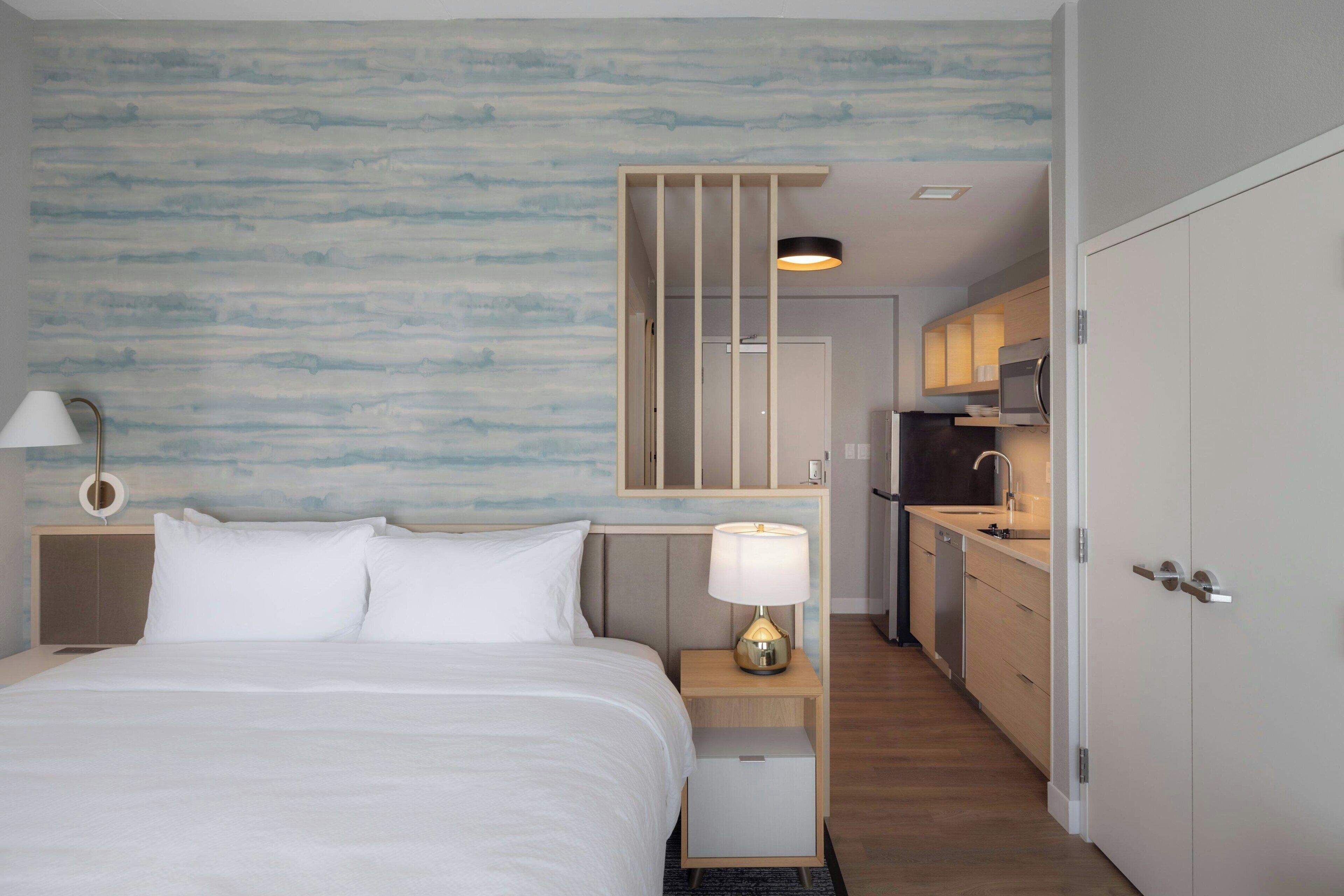 Habitación de hotel con cama, mesita de noche, lámpara y vista hacia la zona de la cocina.