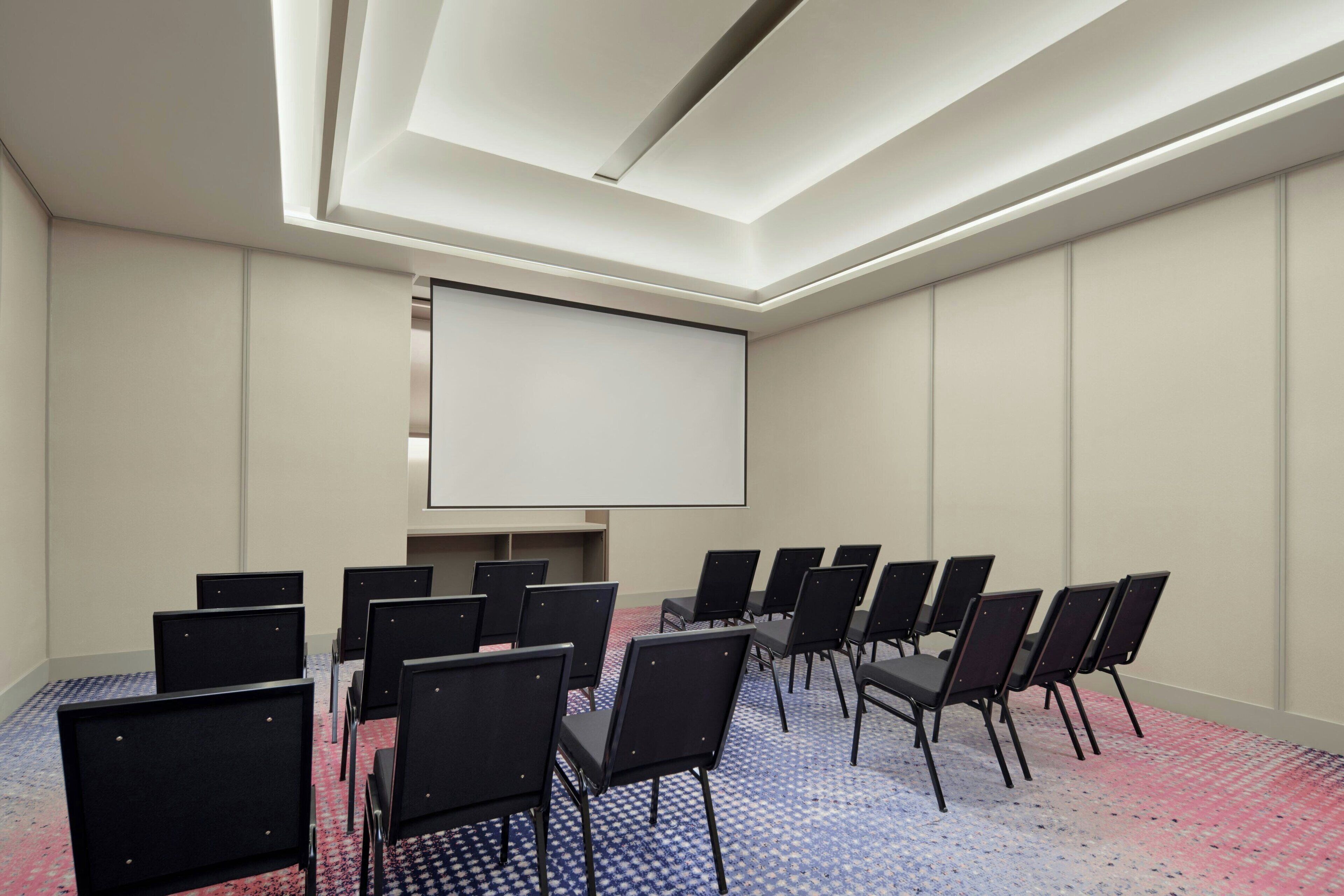 A conference room with a large screen, rows of chairs, and a patterned carpet.