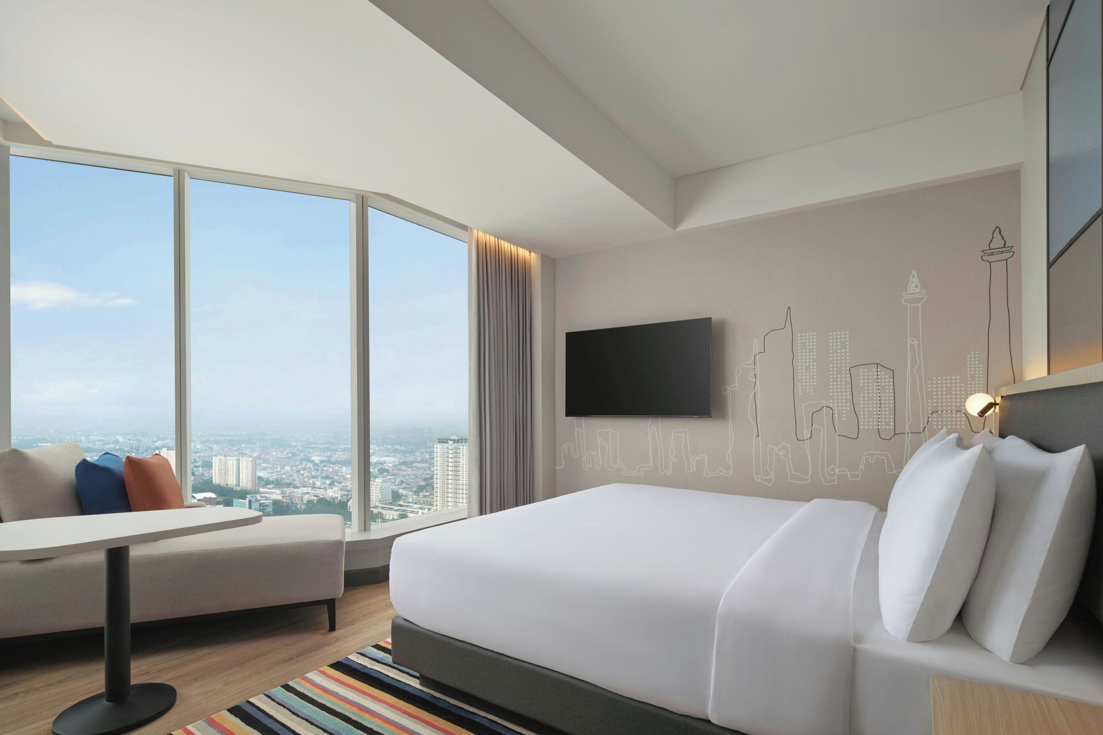 A modern hotel room with a large bed, a sofa, a flat-screen TV, and a cityscape view through large windows.