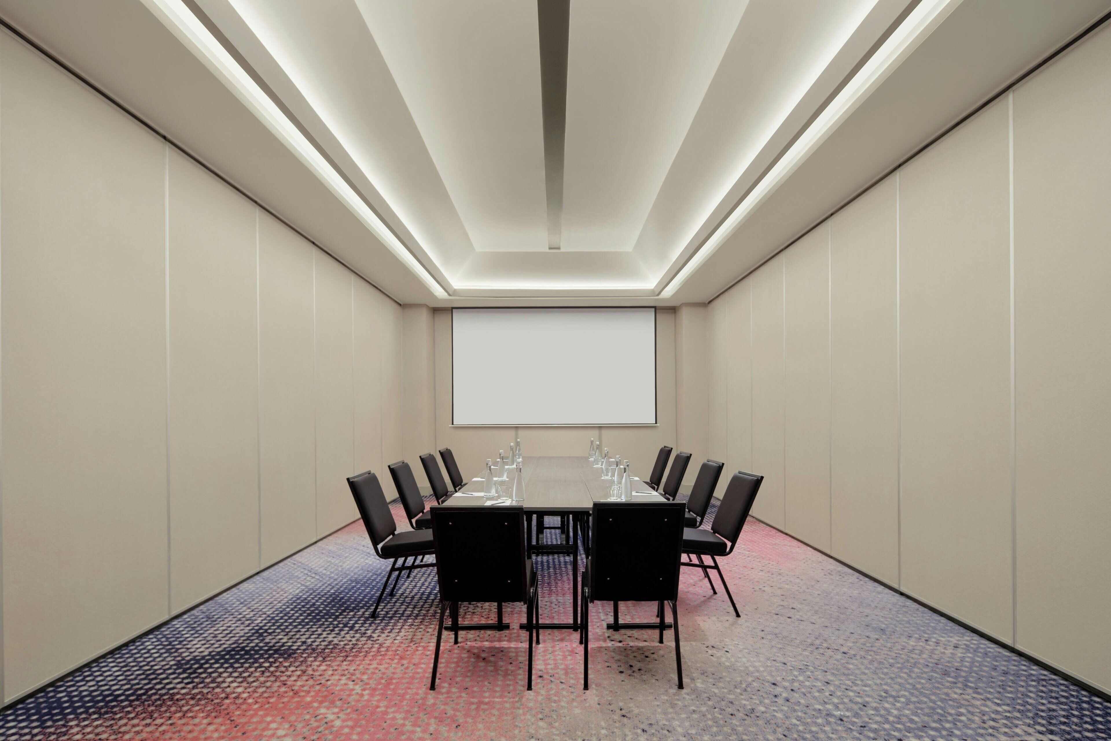 A conference room with a long table, black chairs, and a projector screen.