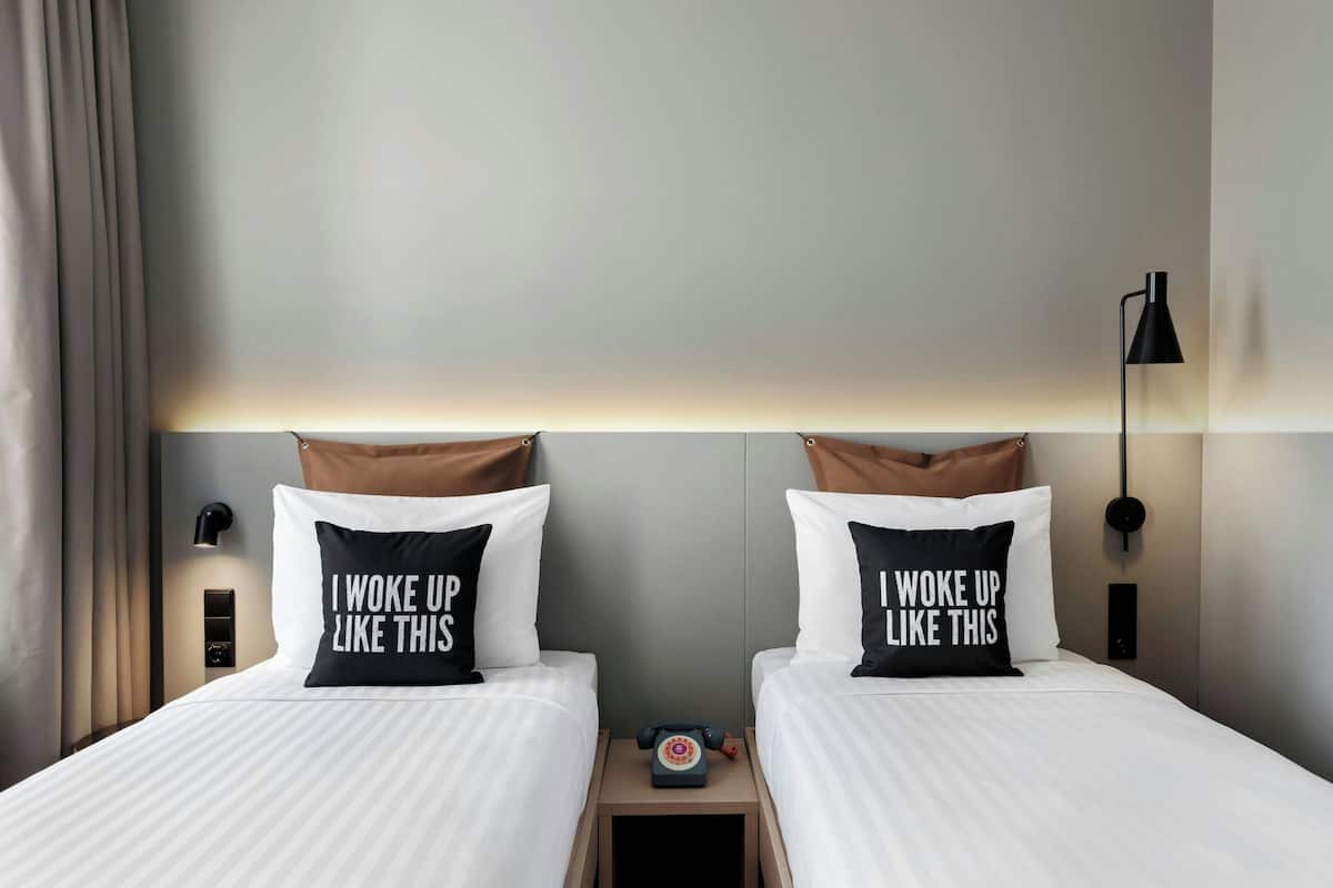 A hotel room with two beds, each with a pillow that says "I woke up like this." There is a nightstand with a lamp and a telephone between the beds.