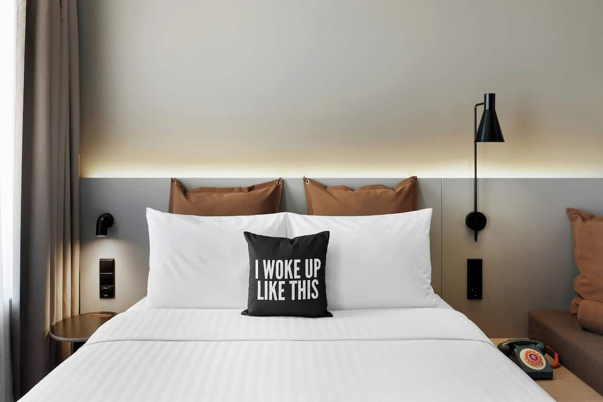 A hotel room with a bed, a pillow with text, a bedside table, a lamp, and a wall-mounted phone.