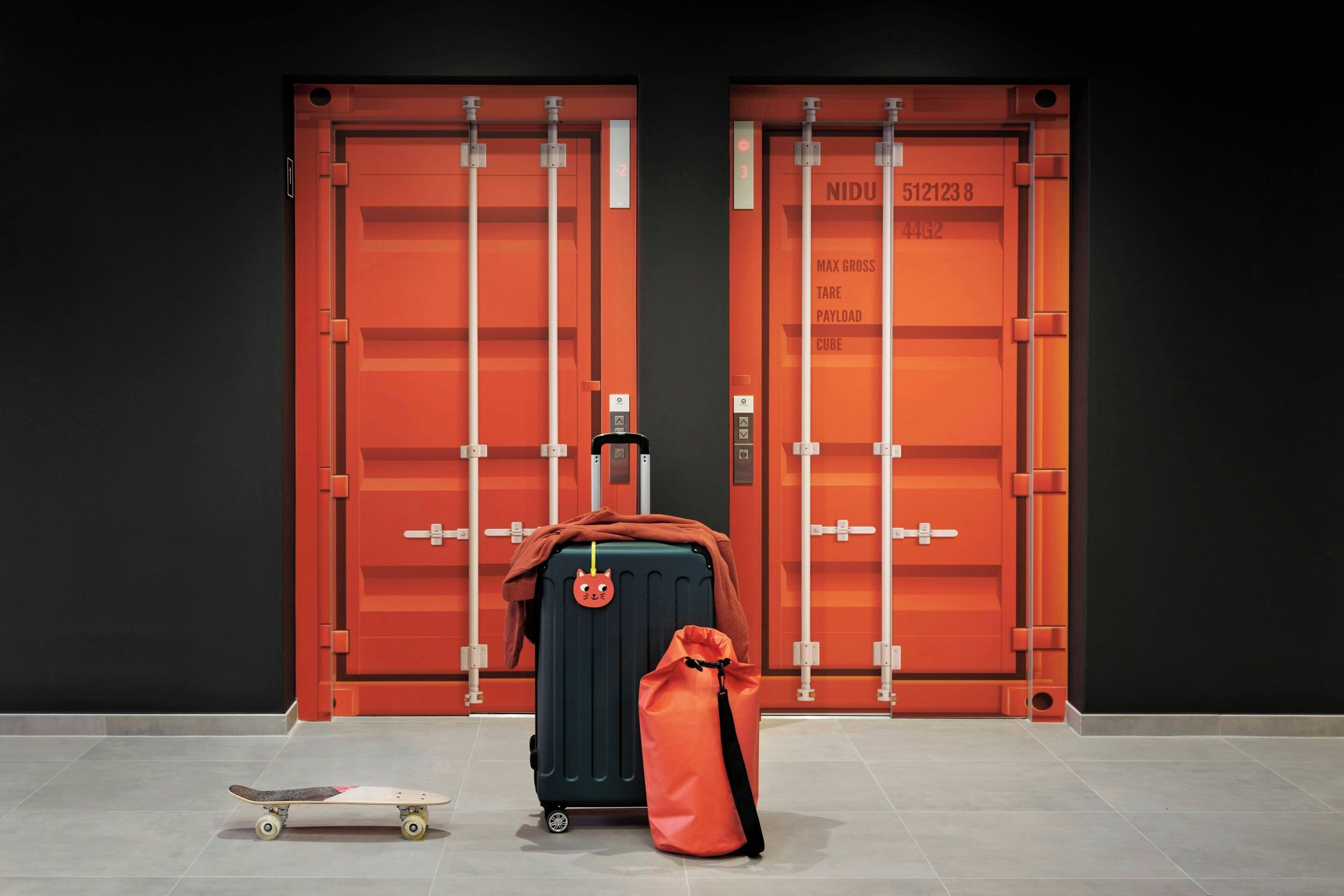 Une valise à la couverture rouge et un petit sac devant une porte rouge ornée d’une plaque avec un numéro.