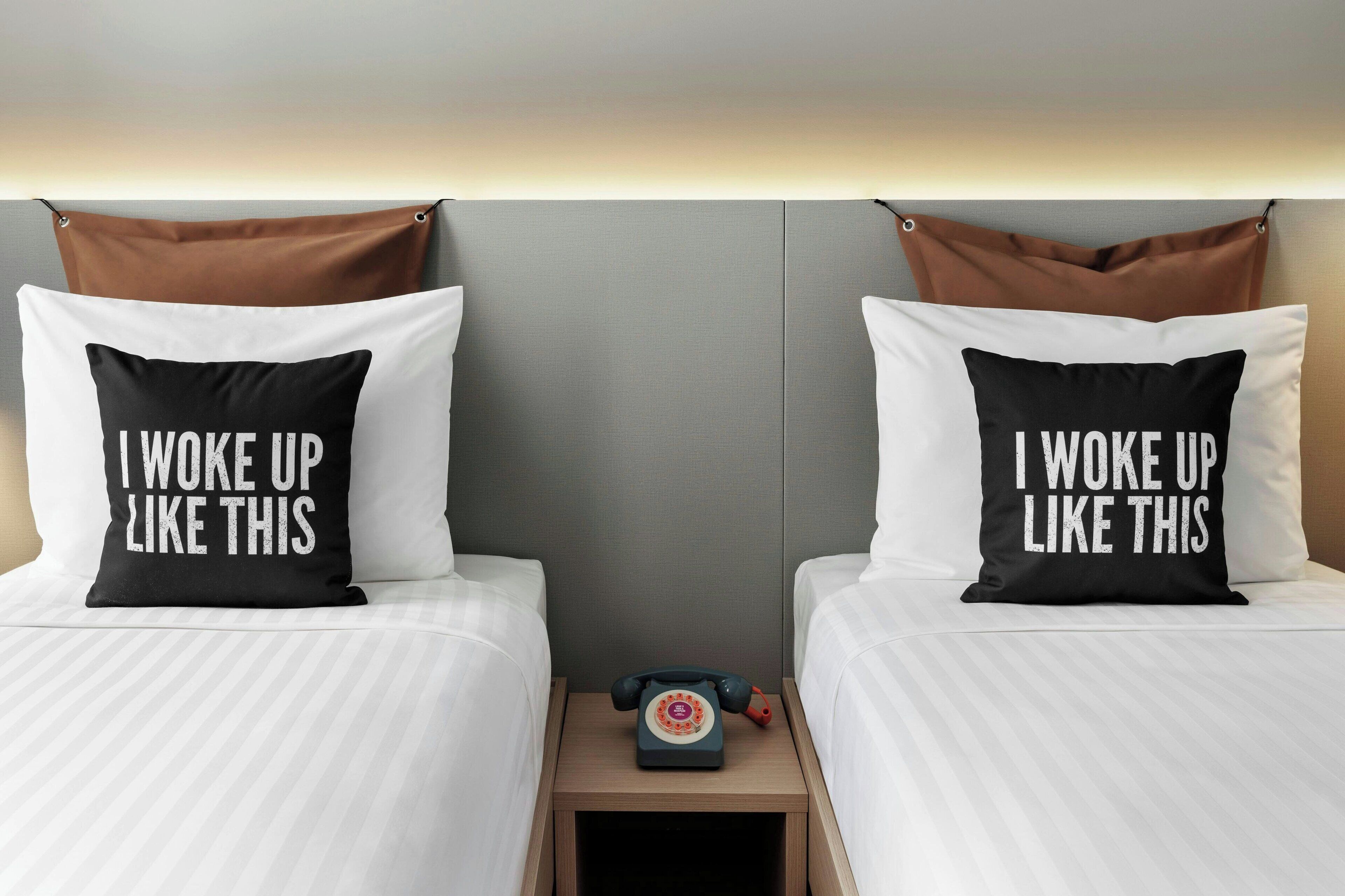 Ein Hotelzimmer mit zwei Betten, jedes mit einem Kopfkissen, auf dem 'I woke up like this' steht. Auf einem hölzernen Nachttisch zwischen den Betten befindet sich ein altes Telefon mit Wählscheibe.