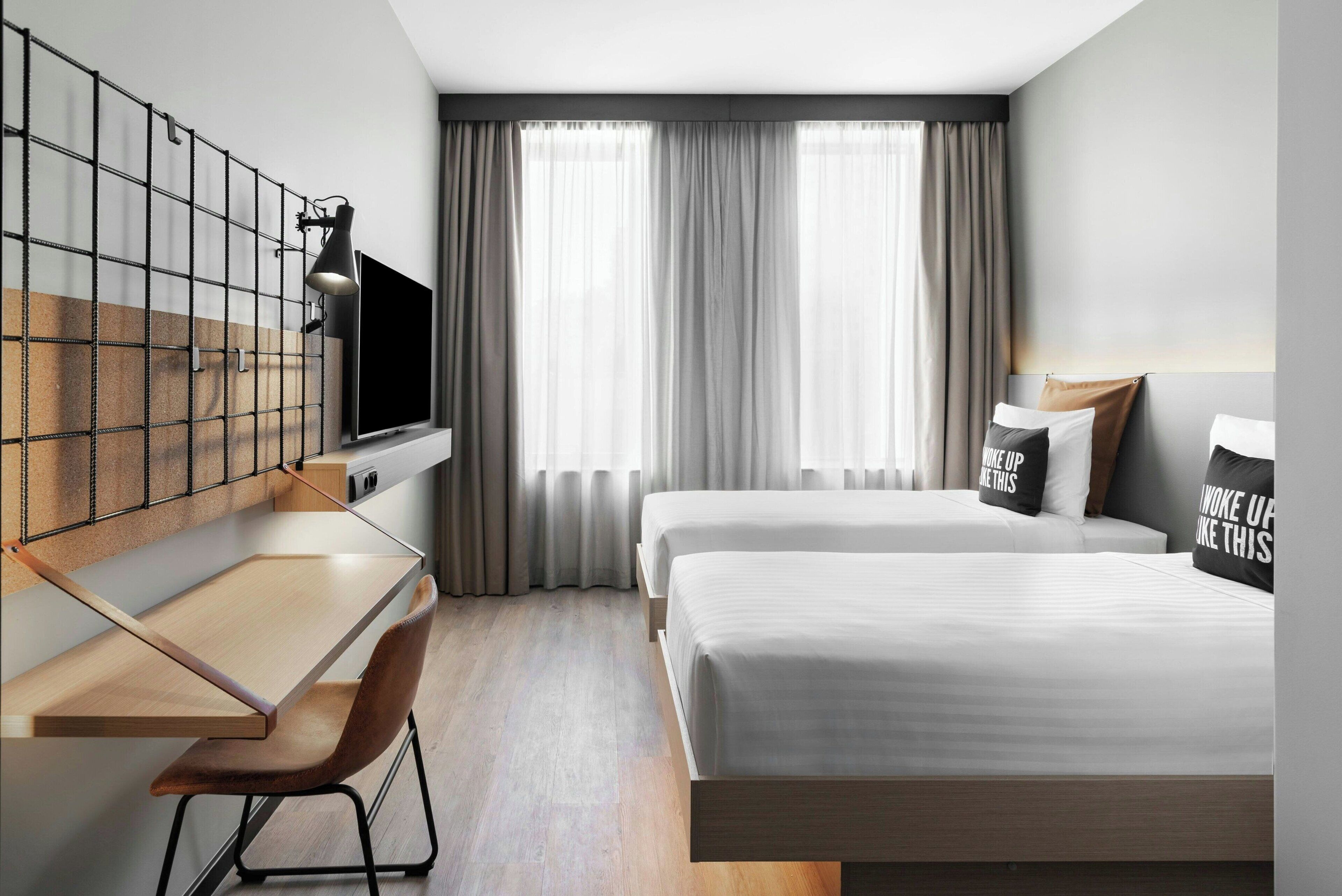 Una habitación de hotel moderna con escritorio, dos camas, televisor y un ventanal con cortinas.