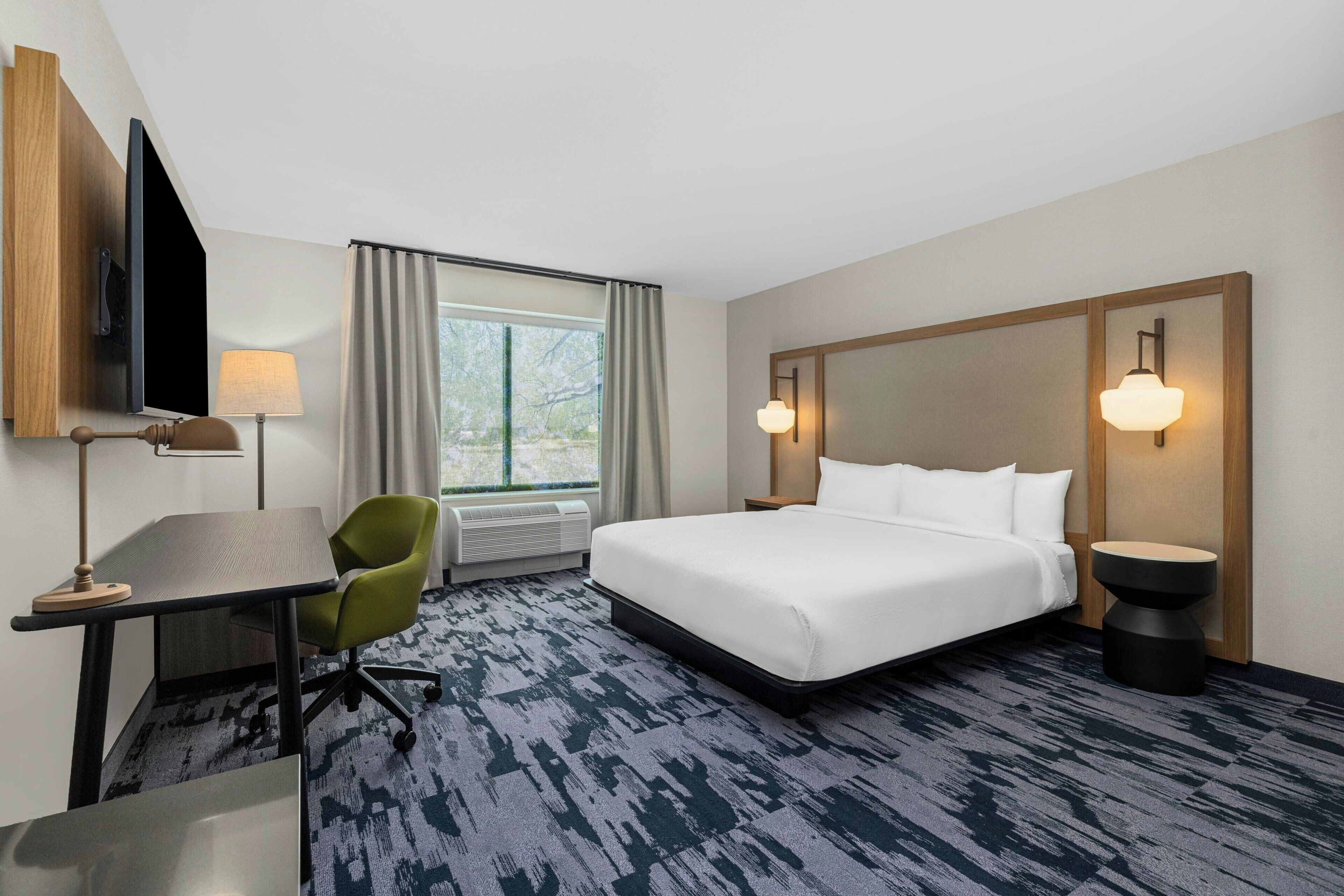 Una habitación de hotel con cama, escritorio, silla, televisión y ventana con cortinas.