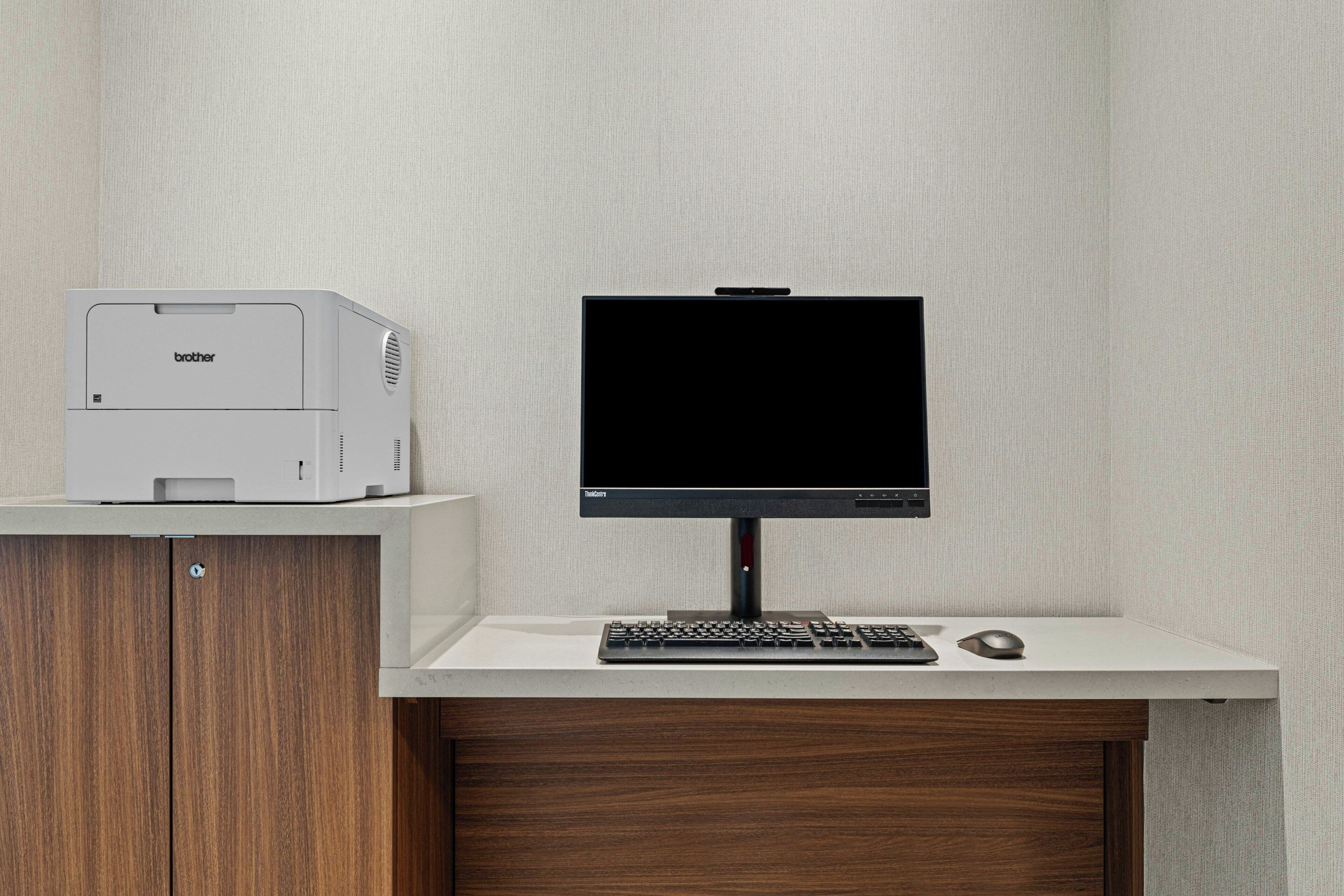 A modern office setup with a Brother printer, a flat-screen monitor, a keyboard, and a mouse on a white desk against a textured wall.