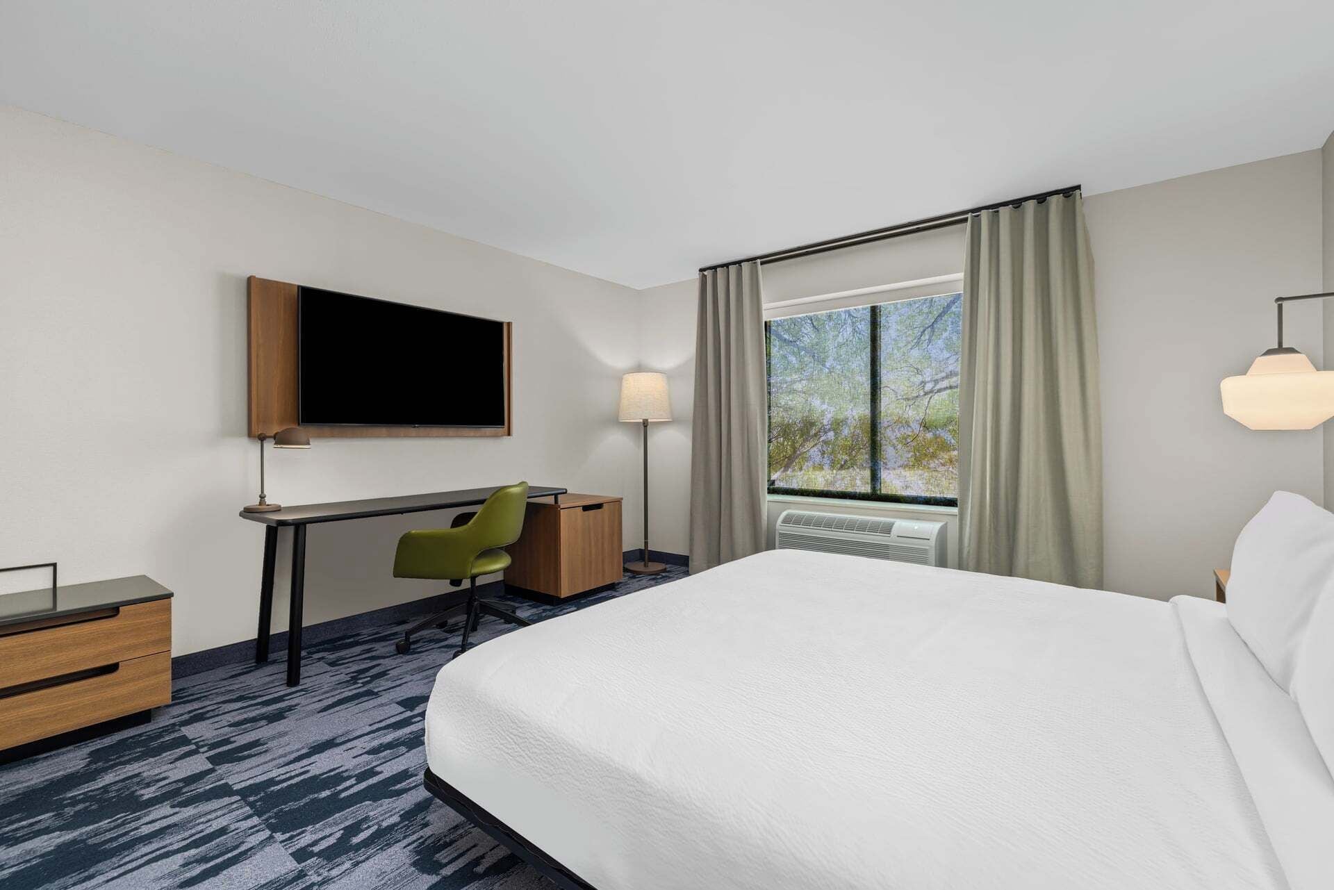 A hotel room with a large bed, a desk with a chair, a flat-screen TV, a window with curtains, and a ceiling lamp.