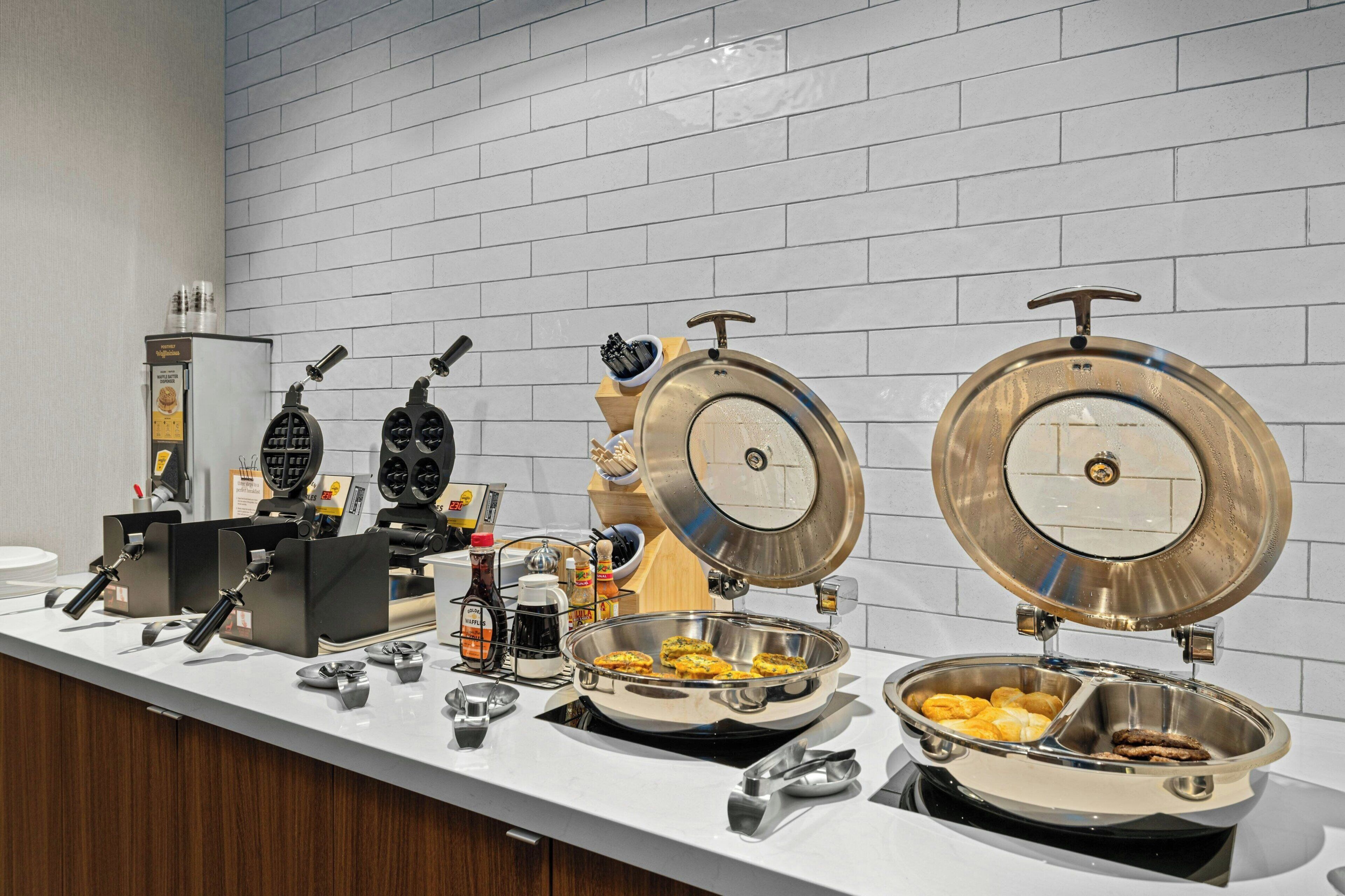 A breakfast buffet with waffle irons, coffee machines, and various food items.