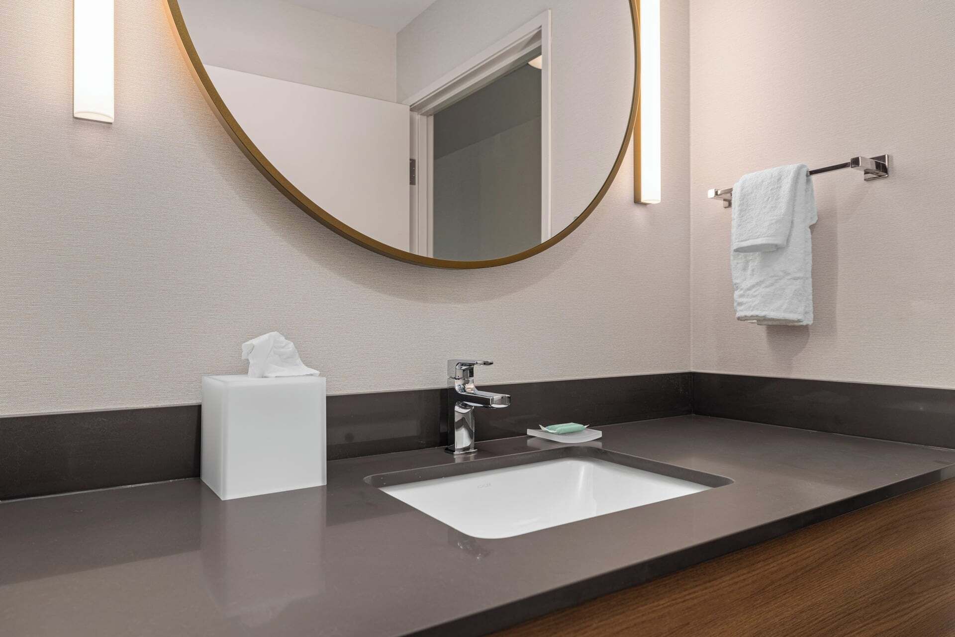 Un baño moderno con un lavabo blanco, grifo cromado y un espejo redondo con marco dorado.
