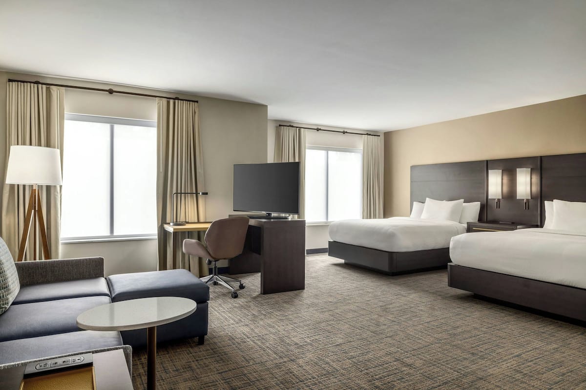 A hotel room with a sofa, a desk with a computer, a TV, two beds, and large windows.