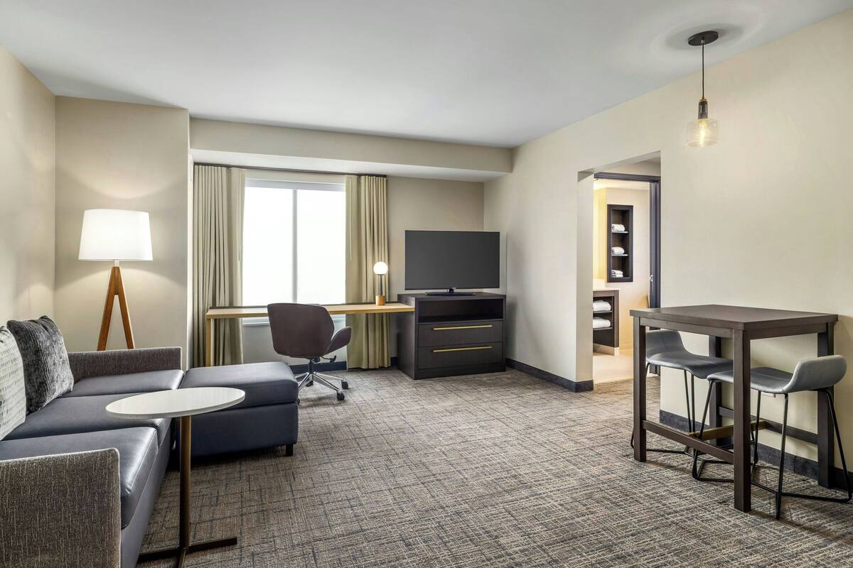 A hotel room with a sofa, a small round table, a desk with a TV, a chair, and a bar area with stools.