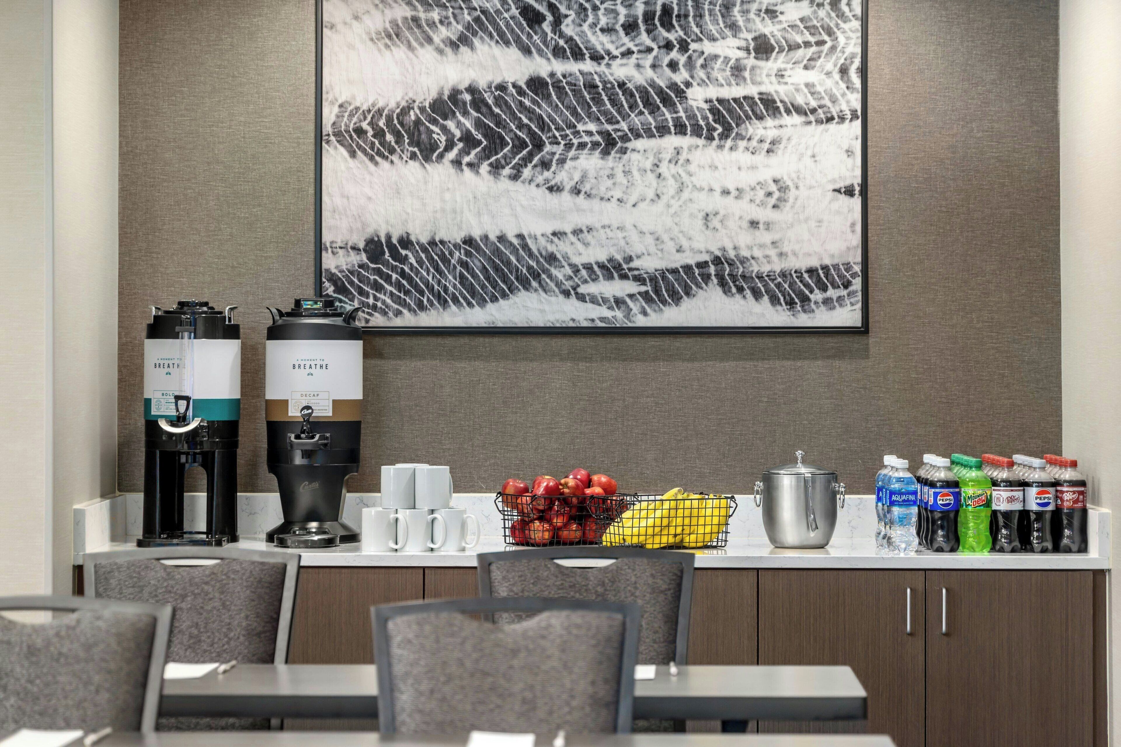 Un comptoir de petit-déjeuner avec des machines à café, un panier de fruits et des boissons en bouteille.