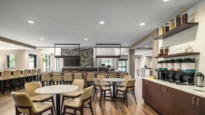 Ein moderner Hotel-Speisebereich mit runden Tischen und Stühlen, einer Küchenzeile mit Kaffeemaschinen und einem großen Fernsehbildschirm.