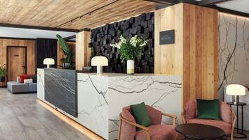 A reception desk with a marble top, wooden paneling, and a black wall with a plant. There are two chairs and a table in the foreground.