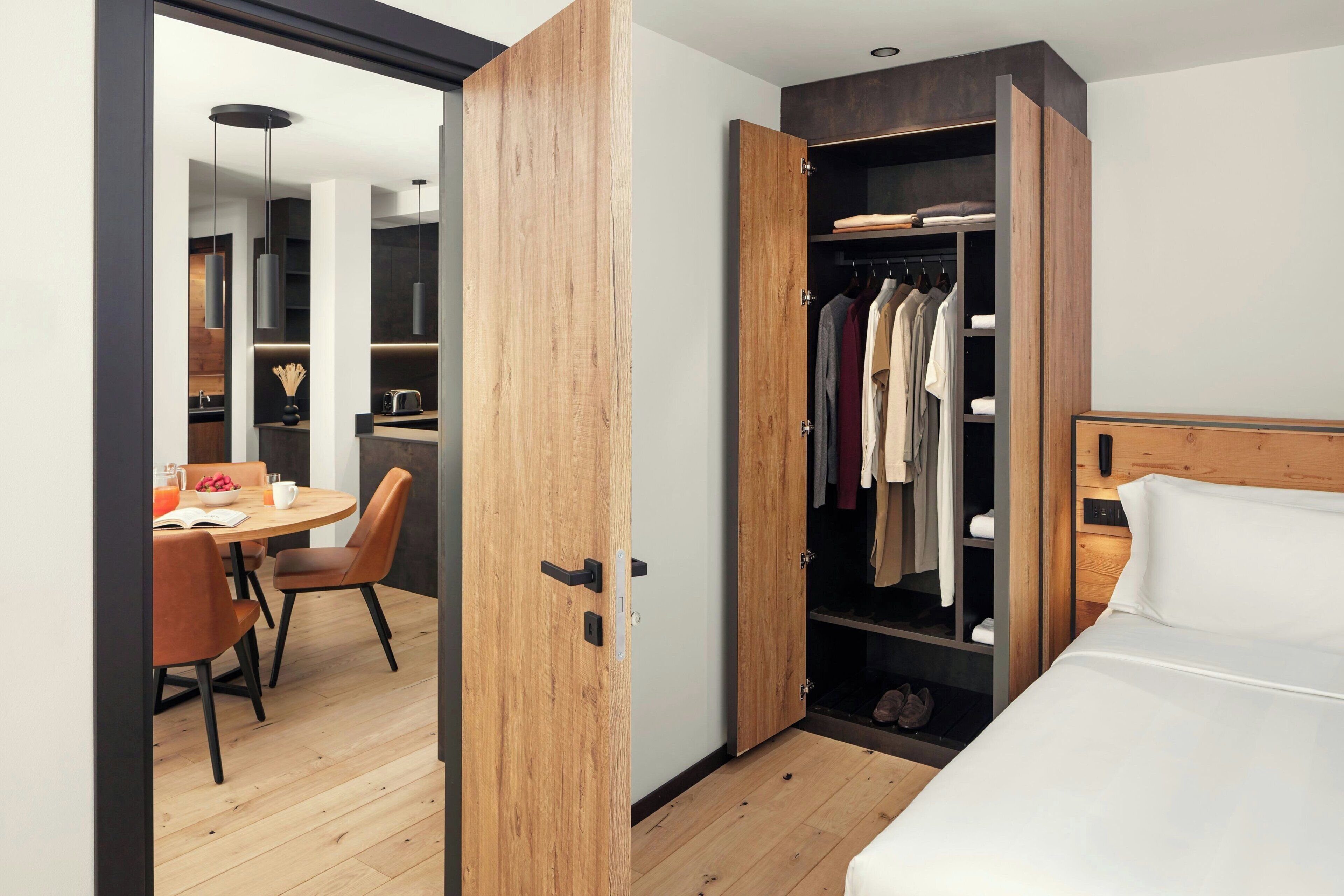 Una habitación de hotel moderna con un armario de madera, un rincón de comedor con una mesa y sillas, y una cama con ropa de cama blanca.