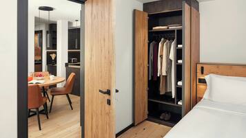 Una habitación de hotel moderna con un armario de madera, un rincón de comedor con una mesa y sillas, y una cama con ropa de cama blanca.